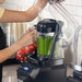 Hamilton Beach HBH855 Summit blender in use, hands pouring a green smoothie with the sound enclosure raised open