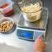 San Jamar Escali SCDGM66 portioning scale in use, hand pressing the keypad while weighing shredded cheese in a pitcher
