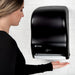San Jamar T1400TBK black touchless roll paper towel dispenser mounted on a wall, towel dispensing to a woman's hand