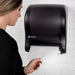 San Jamar T8000TBK black compact touchless paper towel dispenser on a tiled wall, woman tearing off a white paper towel
