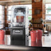 Vitamix 036019-ABAB The Quiet One enclosure blender on a cafe counter beside cups of red smoothies, in use setting