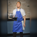 Winco full-length bib apron with pockets in royal blue, worn by a server carrying a tray of drinks in a restaurant