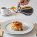 Winco glass syrup dispenser shown in use pouring maple syrup over a stack of pancakes at a breakfast table