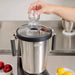 Hamilton Beach HBF1100SR Expeditor 4L blender shown in use as a hand removes the lid cap to add ingredients