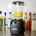 Hamilton Beach HBH650R The Tempest 64 oz blender blending a yellow drink on a bar counter with bottles behind