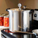 LEM aluminum pressure canner in use on a stovetop, shown in a kitchen beside jars of home-canned tomatoes