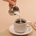 Winco stainless steel bell creamer in use, pouring cream into a cup of coffee on a white saucer