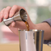 Winco stainless steel jigger shown in use, pouring a measured shot of liquor into a stainless steel bar shaker tin